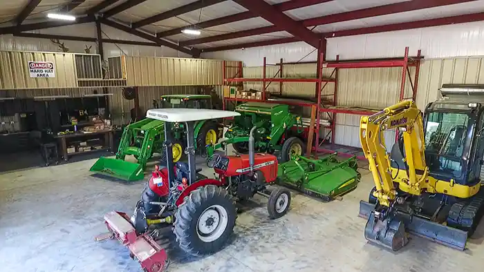 Red and green tractors, a rotary mower attachment, and a yellow Komatsu mini excavator inside a metal workshop
