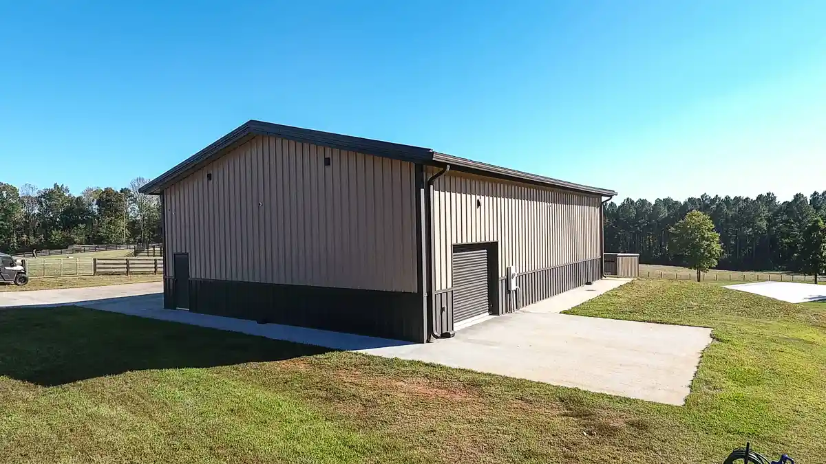 Tan metal building with dark trim, roll-up garage door, concrete driveway, and surrounding grass and trees