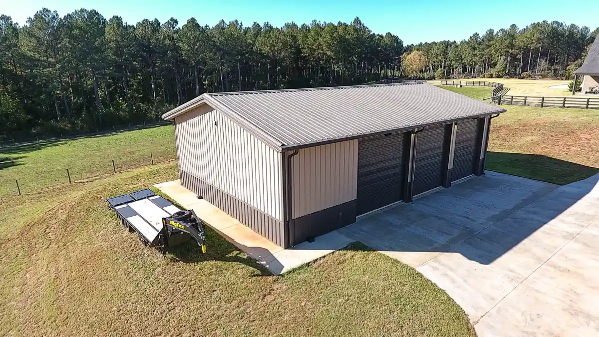 Aerial view of a metal garage building with three bay doors, concrete driveway, and a flatbed trailer parked beside it near a wooded area