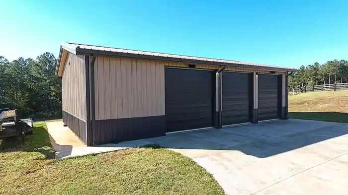 Tan pre-engineered metal building with three dark brown garage doors and a concrete driveway, surrounded by trees