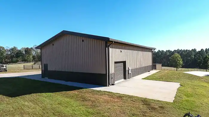 Tan metal building with dark trim, roll-up garage door, concrete driveway, and surrounding grass and trees