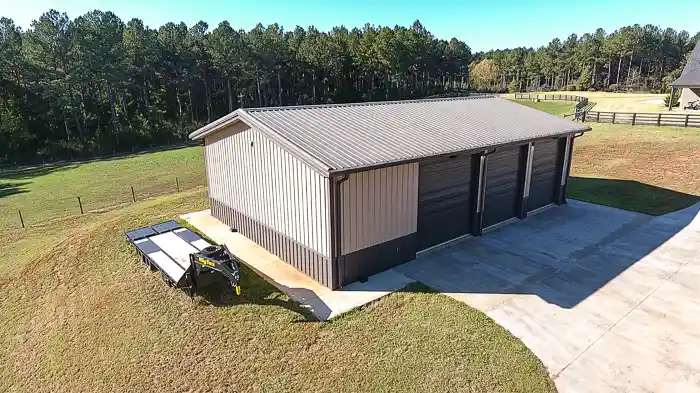 Aerial view of a metal garage building with three bay doors, concrete driveway, and a flatbed trailer parked beside it near a wooded area